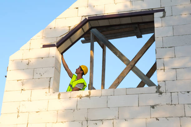 istock Construction worker at construction site measures the length of the window opening and the wall with tape measure. Cottage are made of porous concrete blocks, protective clothing - hardhat and a vest 1332793728
