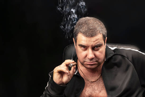 male portrait, a man Smoking a cigar. thug, experienced criminal Smoking a cigar sitting on a chair. Russian mafia. Plowman's in jail, don. copy space, black and gloomy background stock photo
