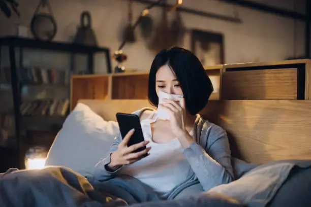 Photo of Young Asian woman suffering from cold and flu, blowing her nose with tissue while using smartphone on the bed and taking a rest at home