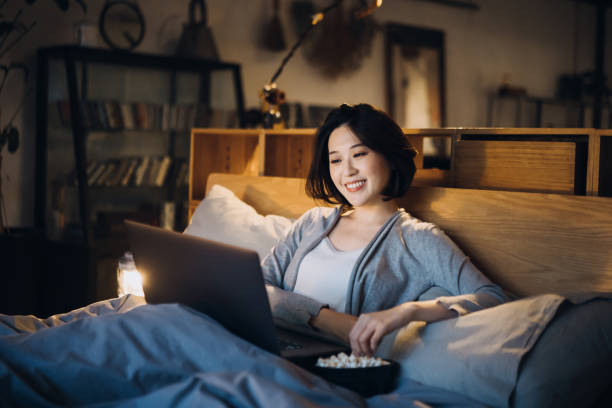 cheerful young asian woman lying on bed enjoying the weekend, watching movie on laptop and eating popcorn at home in the evening - lying down women laptop freedom imagens e fotografias de stock