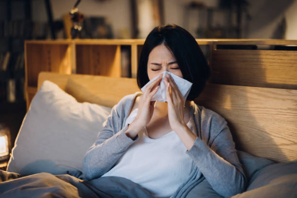 istock Young Asian woman suffering from cold and flu, blowing her nose with tissue while lying on bed and taking a rest at home 1332798292
