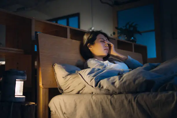 Young Asian woman feeling sick and suffering from a headache, lying on bed and taking a rest at home