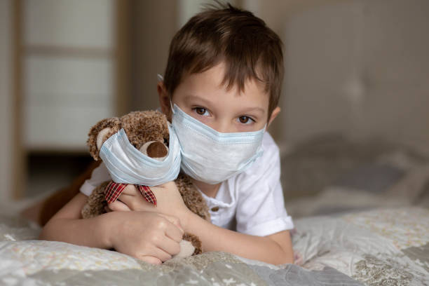 istock A boy in a mask laying on the bed and hugging a teddy bear in a mask. Image with selective focus 1332822714