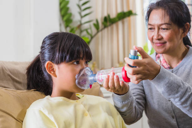 asiatische mutter hilft ihrer tochter, hilfe mit asthma-inhalator spacer zu erhalten, während sie sich auf ein sofa im wohnzimmer zu hause setzt - asthmainhalator stock-fotos und bilder
