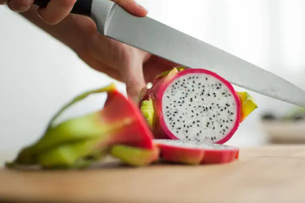 Photo of Female hands is cutting a dragon fruit or pitaya with pink skin and white pulp with black seeds on wooden cut board on the table. Exotic fruits, healthy eating concept