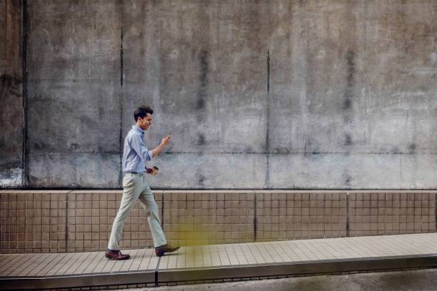 sorridente giovane uomo d'affari asiatico in abbigliamento casual utilizzando il telefono cellulare mentre cammina vicino al muro dell'edificio urbano - city wide foto e immagini stock