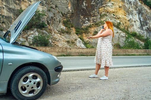 Annoyed overweight woman standing by her broken car on the road and using phone to call for help