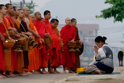 Row of Thai monks walking along people is   getting food donations  at  ceremony of Makha Bucha Day in Sangkhla Buri in northwest of Kanchanaburi province of Thailand in early morning close to sunset. People are wearing mostly traditional clothing and are standing in main street leading down to Mon Bridge and lake. People are waiting to give food donations to monks when they pass along street down to bridge.