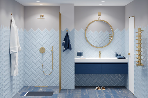 A modern bathroom in blue tones with gold fittings, a bathrobe next to the shower, a round mirror over a large washbasin with a blue cabinet, a golden heated towel rail next to a white door.