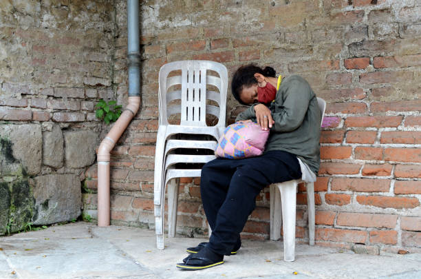 femme sans abri dormant sur une chaise monobloc sur le côté du mur de briques dans un vieux cimetière. - brick wall homelessness wall begging photos et images de collection