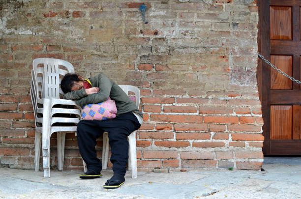 femme sans abri dormant sur une chaise monobloc sur le côté du mur de briques dans un vieux cimetière. - brick wall homelessness wall begging photos et images de collection