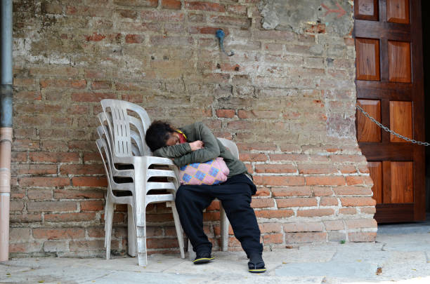 femme sans abri dormant sur une chaise monobloc sur le côté du mur de briques dans un vieux cimetière. - brick wall homelessness wall begging photos et images de collection