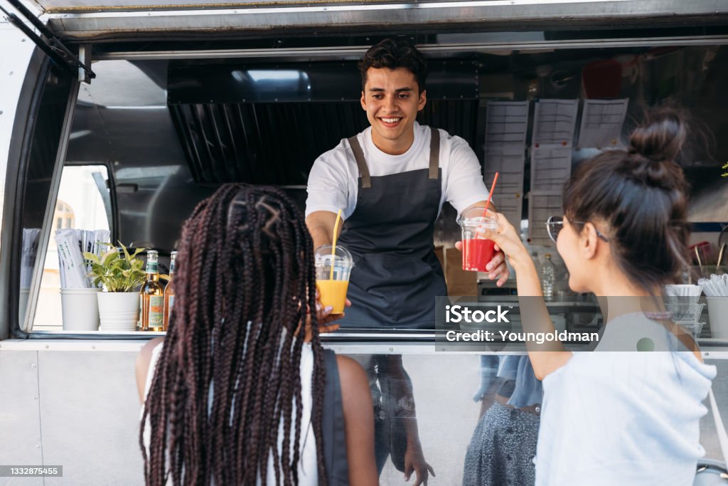 Back view of two females buying drinks from a male owner at a food truck. Smiling entrepreneur giving drinks to clients. Food Truck Stock Photo