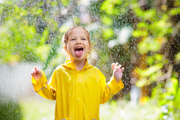 Child playing in the rain on sunny autumn day. Kid under heavy shower with yellow duck umbrella. Little boy with duckling waterproof shoes. Rubber wellies boots. Fall outdoor activity by rainy weather