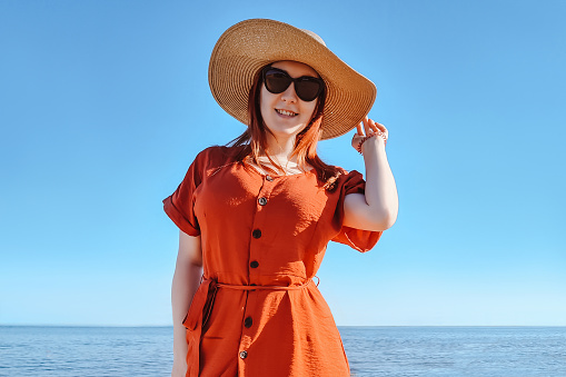 Attractive red-haired woman in sunglasses and a straw hat on the beach. Summer sea vacation. Travel lifestyle.