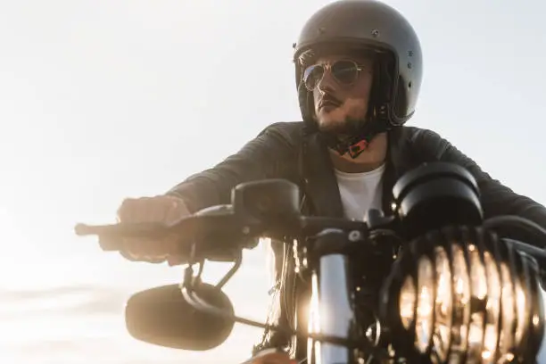 Portrait of biker looking away, sitting on his vintage motorcycle, wearing leather jacket, helmet and sunglasses