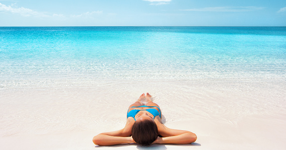 Tropical Caribbean beach vacation - suntan relaxation woman. Bikini girl lying down relaxing on white sand exotic destination. Girl sunbathing during summer holidays or winter getaway.