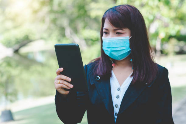 istock business woman wearing face mask using smartphone tablet typing on screen working with app devices in park with sunrise and blur nature background. business new normal with corona virus or covid-19. 1332965347