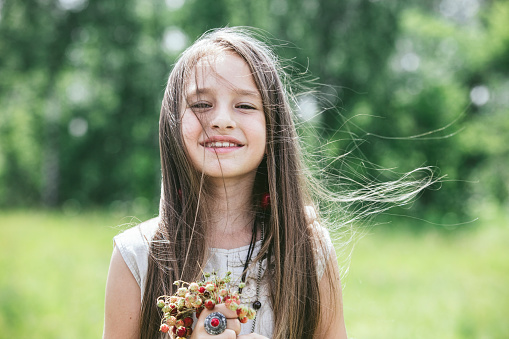 a charming, sweet, little girl with beautiful brown eyes, in a white dress, smiling, dressed in a hippie style, hugging her shoulders, against a background of green grass.