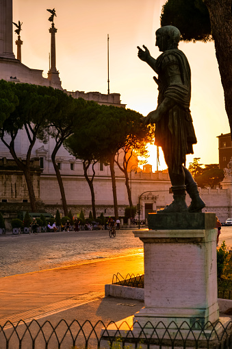 A stunning sunset light envelops the silhouette of the statue of the emperor Trajan along the Fori Imperiali boulevard, in the ancient heart of Rome. This bronze statue, placed in front of the Trajan's Forum, is a copy of the original marble preserved in the National Archaeological Museum of Naples, made in the first century AD. and found in the city of Minturno during some archaeological excavations. In the background the silhouette of the National Monument of the Altare della Patria, in Piazza Venezia square. The Altare della Patria is the scene of all the official Italian civic celebrations, in particular the Republic Day on June 2 and the Liberation Day on April 25. In 1980 the historic center of Rome was declared a World Heritage Site by Unesco. Image in high definition format.