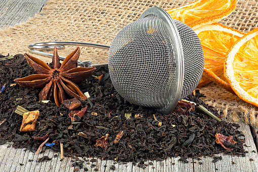 Tea strainer with aromatic tea and dried orange slices on wooden table. Pile of black tea with dried fruits and petals.