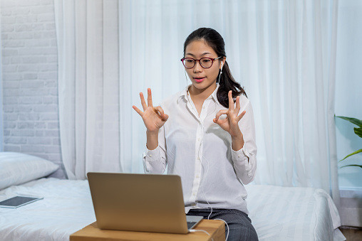 Young asian woman wears headphones to talk and video conferencing on a laptop computer in the bedroom at home.