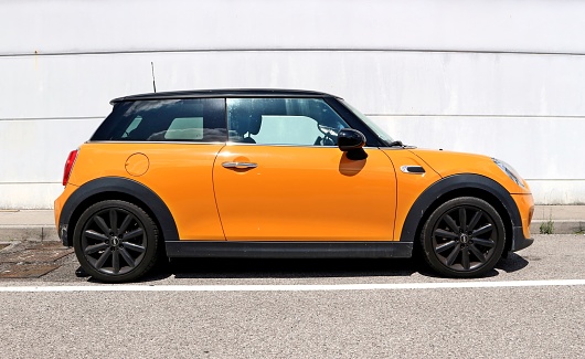 Udine, Italy. August , 6 2021. Side view of an orange Mini Cooper, or Mini Hatch, parked on the roadside. Concrete wall on background.