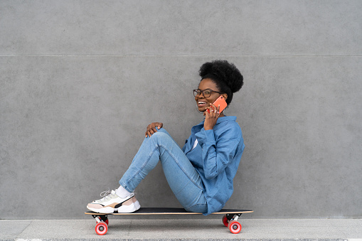 Joyful trendy black girl talking on phone call. Stylish african woman in urban denim outfit calling and communicating using cell phone outdoors. Mobile connection, technology and lifestyle concept