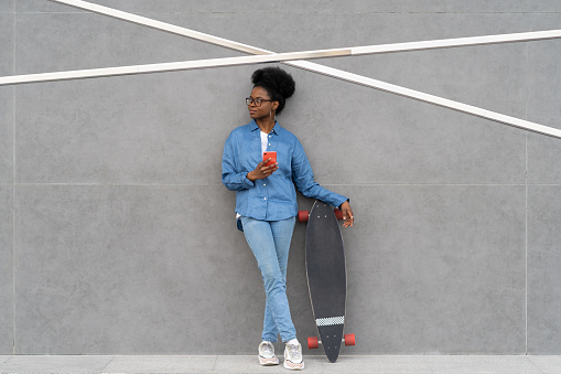 Stylish african american girl with smartphone in hand hold longboard. Trendy casual black woman in denim shirt chat in mobile phone app using 5g internet outdoors after skateboarding. Urban lifestyle