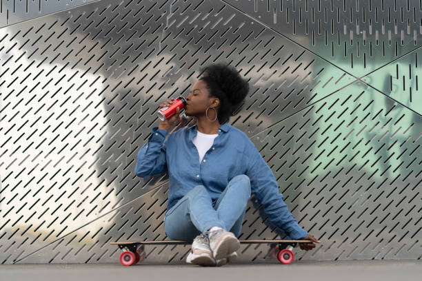 Tired afro american skateboarder girl refreshing after longboarding sit on skate drink soda beverage Tired african skateboarder girl refreshing after longboarding sit on skate drinking soda beverage from metal can. Trendy urban young female relaxing after active training on longboard in urban space non alcoholic beverage stock pictures, royalty-free photos & images