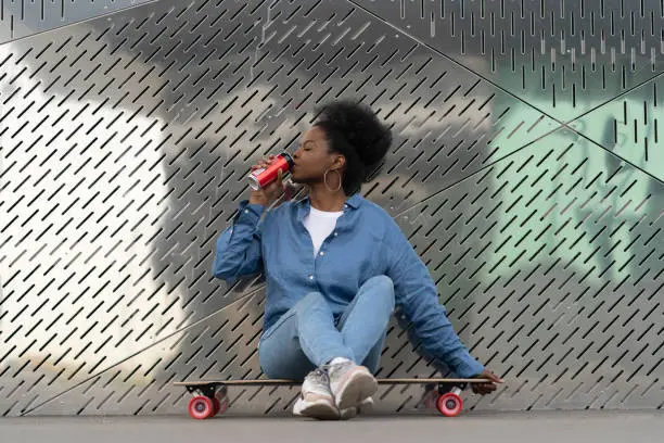 Tired african skateboarder girl refreshing after longboarding sit on skate drinking soda beverage from metal can. Trendy urban young female relaxing after active training on longboard in urban space