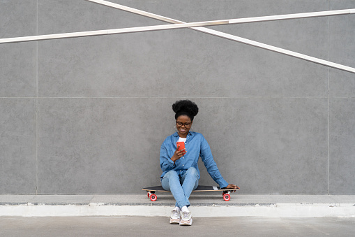 Excited smiling girl texting in smartphone or scrolling social media via 5g internet sitting outdoors on longboard. Trendy african american female with mobile phone in hand over gray concrete wall
