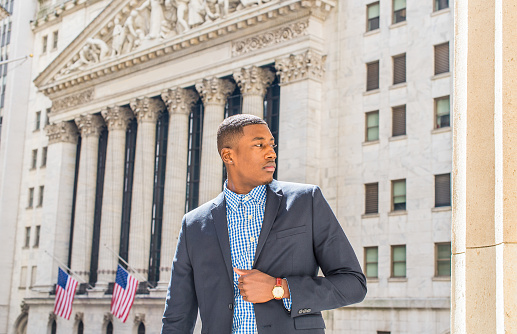 A young black businessman is standing outside an office building, confidently looking forward.\