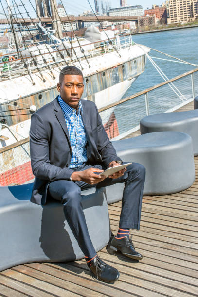 istock Young black man sitting outdoors by river in New York City, reading tablet computer 1333034773
