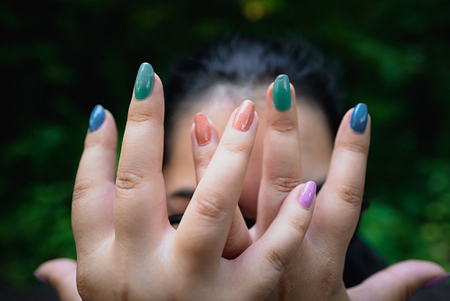 Close up of a woman's hands, with multi colored fingernails, covering her face.
