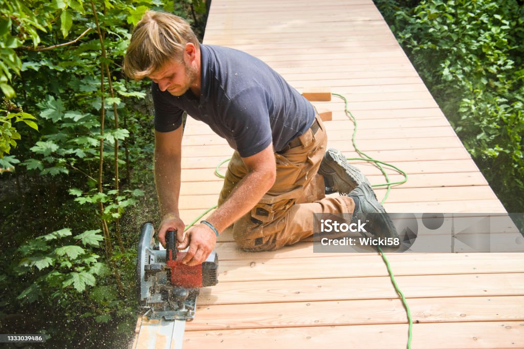 Carpenter straighten edges of boards on a new boardwalk A carpenter cutting off uneven edges of flooring boards with a circular saw, the circular saw is cutting along a metal guide rail to get the edges all even. The new build boardwalk leads over a small river. Power Tool Stock Photo