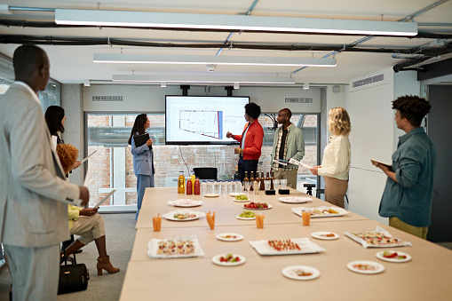 Multi-ethnic group gathering around conference table covered with food and drink in open board room as colleague shares location details on infographic.
