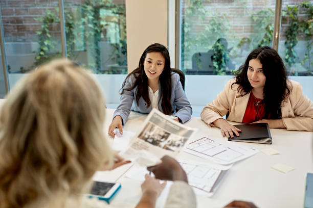 Commercial Real Estate Marketing Team Discussing Property Over the shoulder view of female brokers in 20s and 30s sitting at conference table sharing property information and floor plans. brand strategy stock pictures, royalty-free photos & images