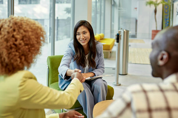 Asian Real Estate Agent Congratulating Clients at Closing Over the shoulder view of young Black couple shaking hands with smiling broker after signing closing documents in modern office lobby. commercial real estate stock pictures, royalty-free photos & images