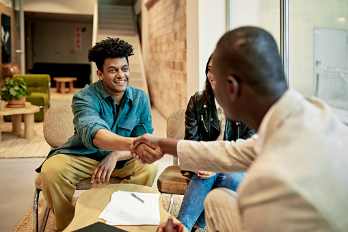 Mid adult couple sitting side by side and smiling as Real Estate Agent congratulates them on their property purchase in modern office lobby.