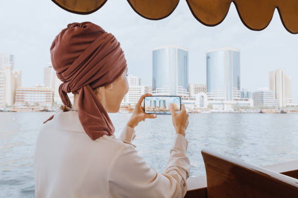 une femme asiatique heureuse dans un turban rouge prend des photos sur son smartphone tout en naviguant sur un bateau traditionnel abra dhow sur la crique de dubaï. voyage aux émirats arabes unis concept - cocklebur photos et images de collection