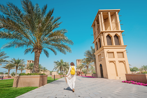 Woman tourist wearing a maroon turban and yellow backpack walks along old wind towers and palms in Bur Dubai and Creek district. Travel and sightseeing spots