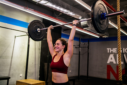 Photo of a fit woman performing a front squat with a heavy barbell in  gym during covid-19 pandemic. Woman lifting a barbel and performing heavy powerlifting exercises. Power clean exercise. Focusing on core strength improvement.