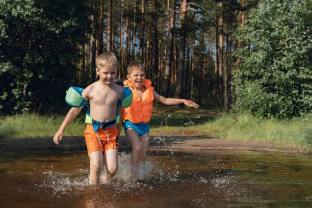 two cute little boys running into lake splashing water in the forest. one boy wearing life vest, another wearing armbands. Pine forest on background. High quality photo