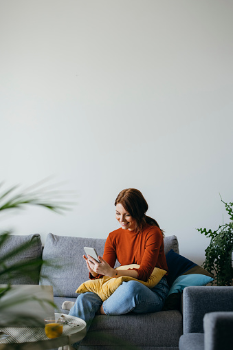 Smiling business woman sitting on couch and typing text message on her smartphone in the living room