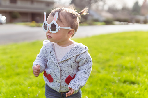 A mixed race one year old little girl walking outdoors on green grass. She's wearing cute bunny sunglasses and has a stoic expression on her face