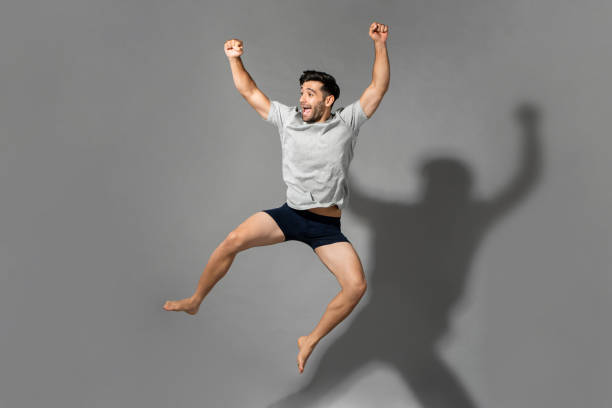 full length portrait of young fresh energetic man wearing sleepwear jumping in mid-air after wake up from a good sleep in the morning - roupa interior imagens e fotografias de stock