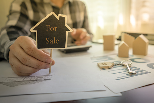 Real estate agents showing message For Sale on the sign of house shape on the table with a calculator, key, house design document, home wooden model.