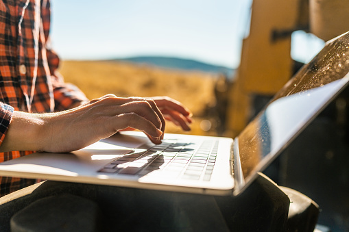 A close-up photo of a female farmer's hands using a laptop next to a combine harvester machine. Smart farming.