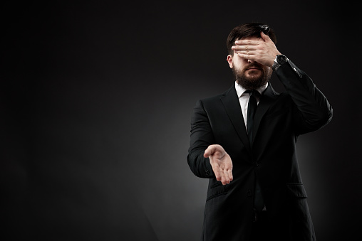 Portrait of bearded businessman in suit and tie standing against black background, demonstrating emotions with gestures. Blind meeting, online business, anonymous deal, secret handshake concept